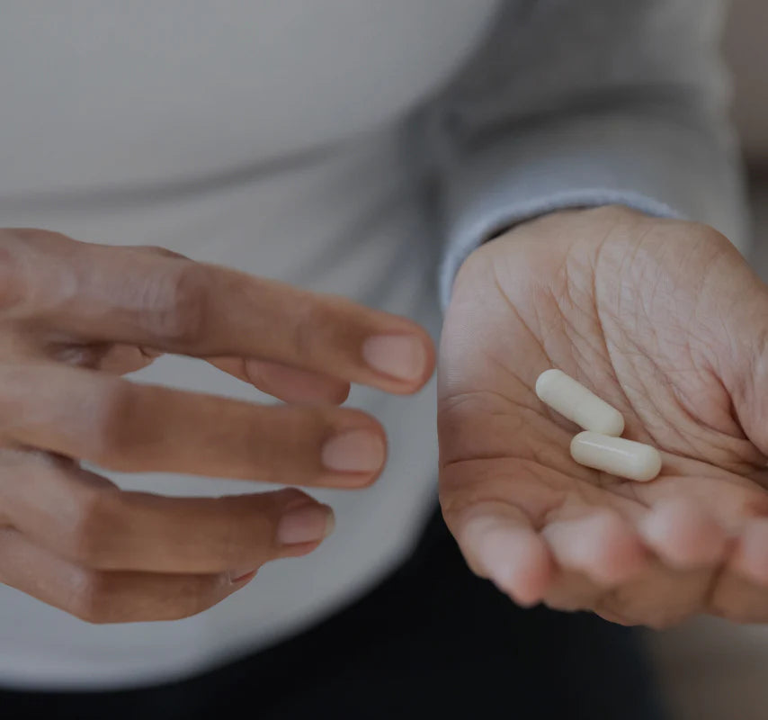 Eine Hand hält zwei weiße Nahrungsergänzungsmittel-Kapseln, während die andere Hand sich an der Nähe der Kapseln befindet. Der Hintergrund ist unscharf, was den Fokus auf die Kapseln und die Hände legt.