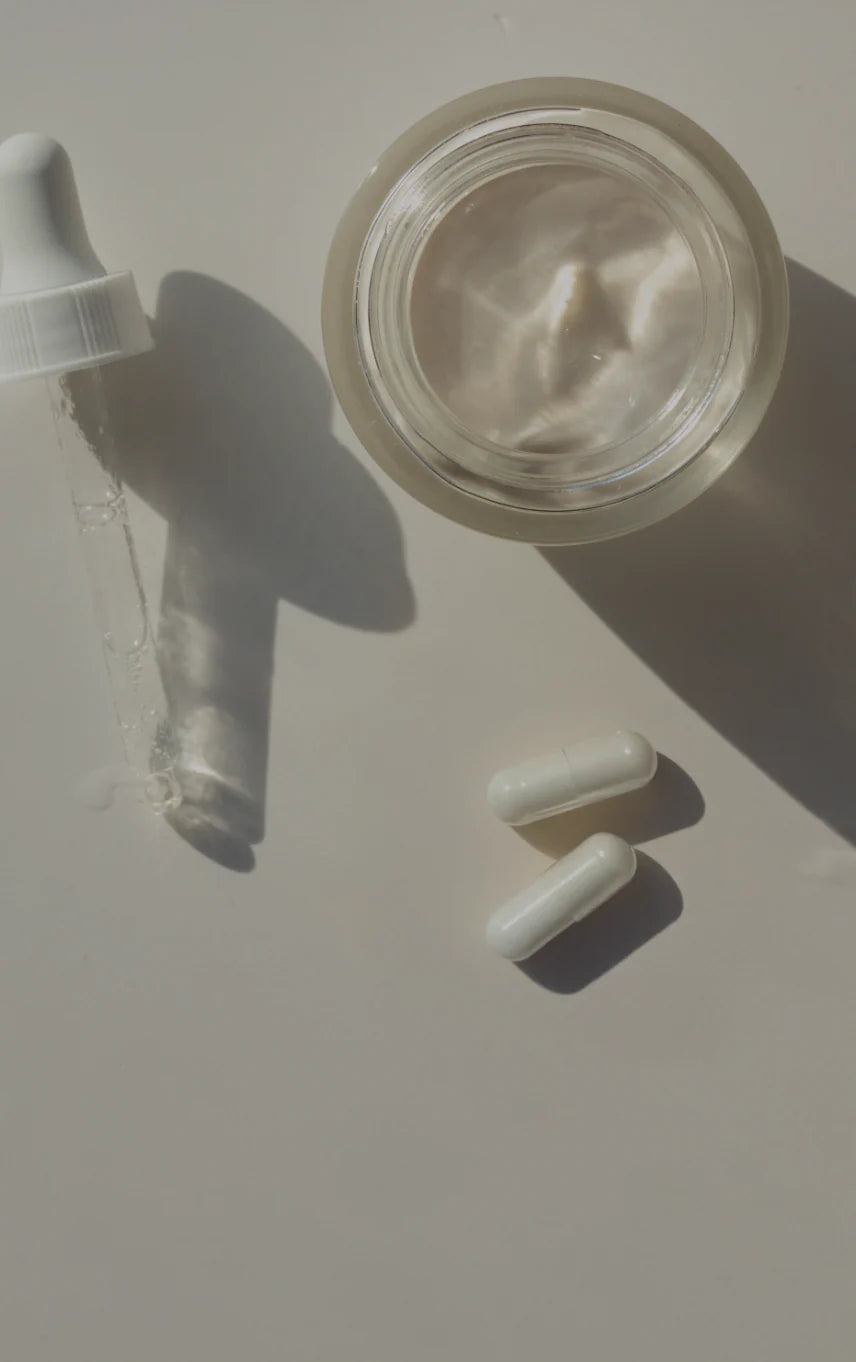 Ein flaches Bild von Nahrungsergänzungsmitteln. Zu sehen sind zwei weiße Kapseln, ein Glasbehälter mit einer klaren Flüssigkeit und ein Tropfer, der neben dem Glas liegt. Die Szene ist gut beleuchtet, mit einem sanften Schattenwurf auf der Unterlage.