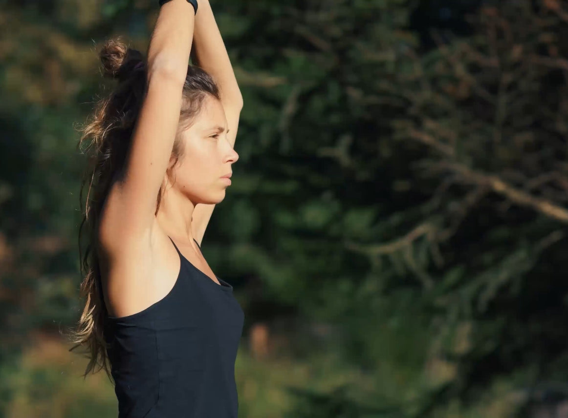 Eine junge Frau in einem schwarzen Tanktop streckt ihre Arme über ihren Kopf. Im Hintergrund sind unscharfe grüne Bäume sichtbar, was eine natürliche Umgebung schafft. Die Frau wirkt selbstbewusst und entspannt.